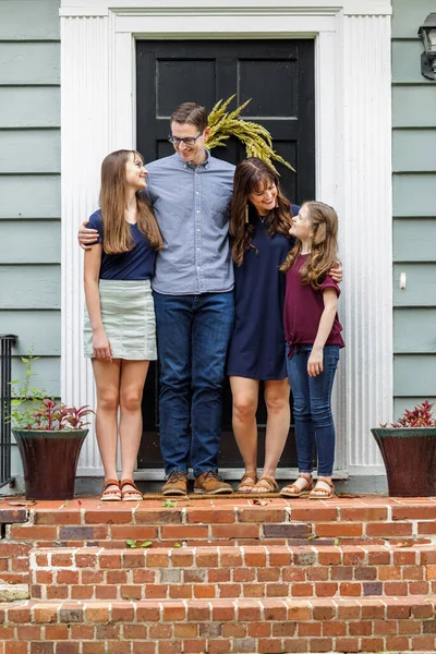 Семья с матерью, отцом и двумя дочерьми, стоящими снаружи на кирпичном крыльце маленького голубого домика, смотрящими друг на друга — стоковое фото