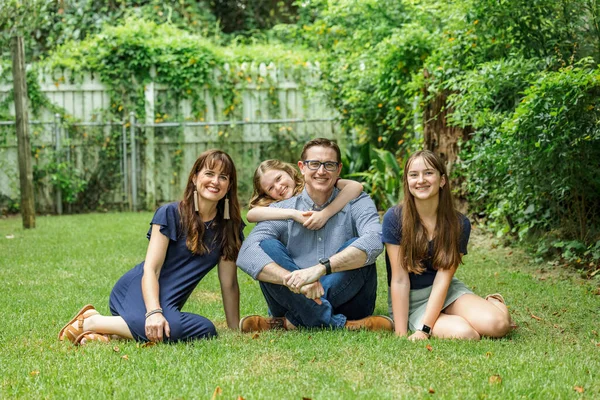 Rodzina matki i ojca i dwie córki siedzące na trawie na podwórku z najmłodszą córką przytulającą tatę od tyłu. — Zdjęcie stockowe