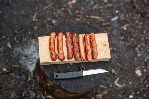 Grilled sausages lie on a wooden board and a knife on a stump. — Stock Photo, Image