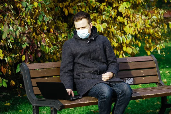 Handsome young European man in a park with laptop with a medical face mask on. Freelance working outside the office during an Covid-19 epidemic. Selective focus