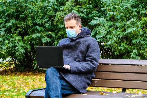 Handsome young European man in a park with laptop with a medical face mask on. Freelance working outside the office during an Covid-19 epidemic. Selective focus