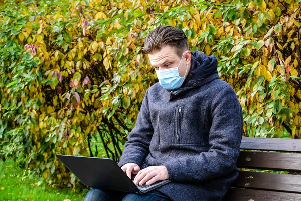 Handsome young European man in a park with laptop with a medical face mask on. Freelance working outside the office during an Covid-19 epidemic. Selective focus