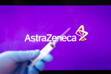 Riga, Letonya: 28 Ocak 2021: AstraZeneca Covid-19 aşı konsepti. Şırınganın yakın çekimi ve arka planda AstraZeneca şirketinin logosu. Odaklanmamış