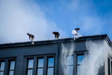 Riga, Letonya: 12 Şubat 2021: Erkek işçiler emniyet kemerlerinde küreklerle kardan binaların çatısını temizledi. Seçici odak