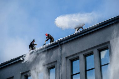 Riga, Letonya: 12 Şubat 2021: Erkek işçiler emniyet kemerlerinde küreklerle kardan binaların çatısını temizledi. Seçici odak