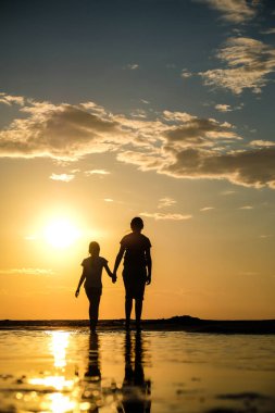 Gün batımında sahilde el ele tutuşan iki kız kardeşin silueti. Dikey fotoğraf