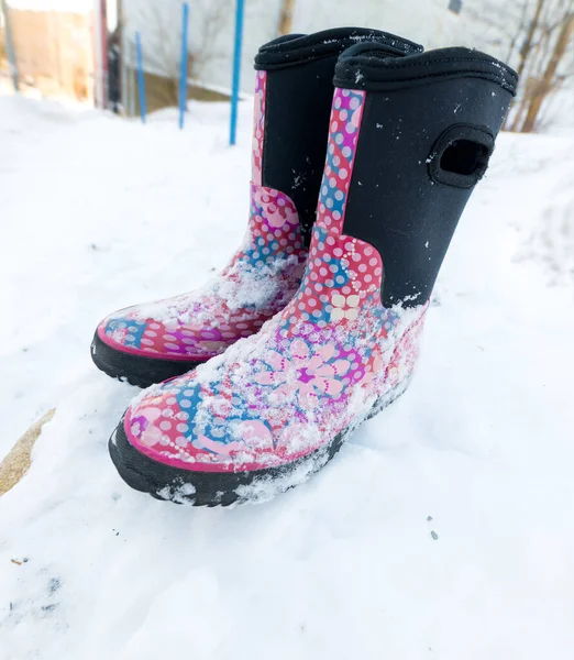 An empty pair of child\'s winter boots resting on a snowbank. The boots are pink, red, blue, and black. There is snow on them.