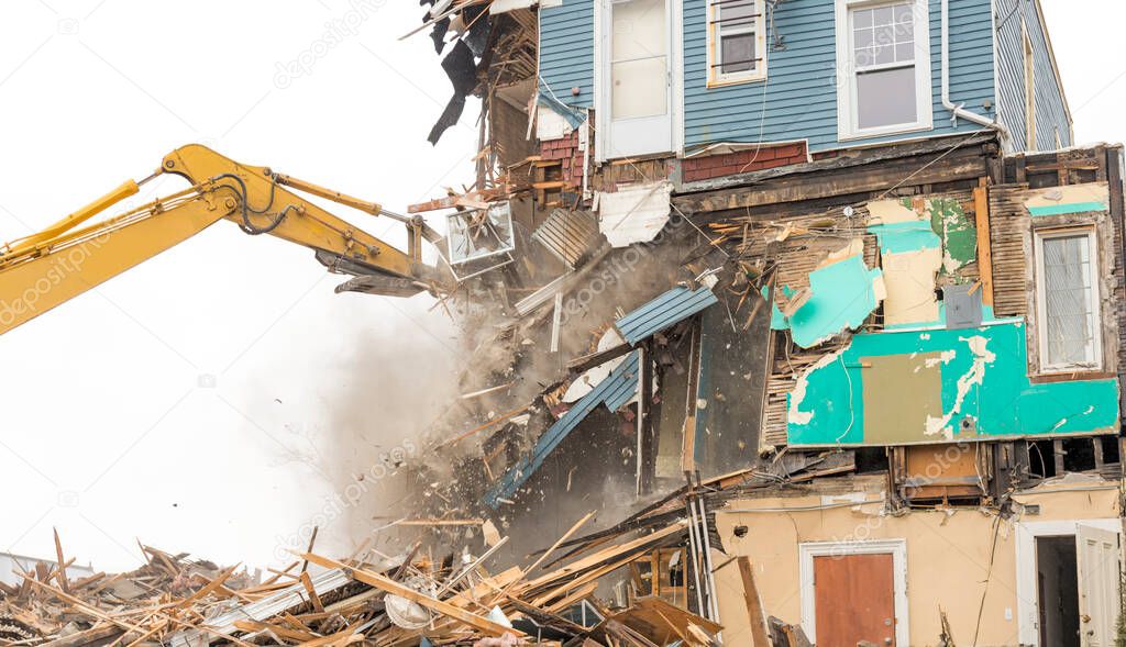 Demolishing a building with a large backhoe. Overcast day. Air is dusty. Lots of debris from the demolition. Much debris falls from a wall being demolished.