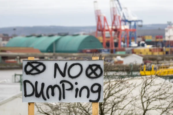 A hand painted no dumping sign. The sign is black letters on a white board, and is held up by wooden posts. Industrial cranes and sheds in the background. Room for text.