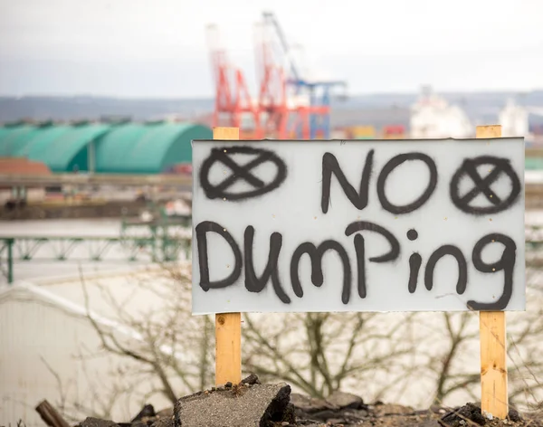 A hand painted no dumping sign. The sign is black letters on a white board, and is held up by wooden posts. Industrial cranes and sheds in the background. Room for text.