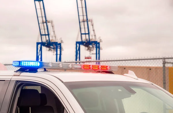 Lights on a police car at a port. The lights red and blue. Only top of car and lights are visible. Cargo cranes and fence are in the background.