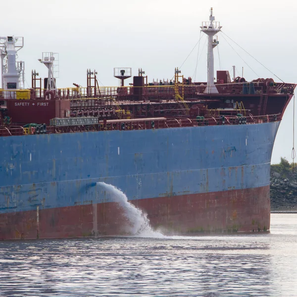 Tanker discharging ballast into the harbor. Water flows from the side. Ship not identifiable. Only part of ship visible. Overcast, hazy day.