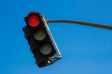 Bir açıdan trafik ışığı, güçlü bir rüzgarla savruluyormuş gibi. Kırmızı ışık yanıp sönüyor. Gökyüzü mavidir.