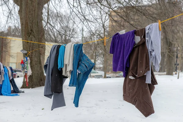 Clothing hanging on a rope in a park. The clothing was left for homeless and other people in need. Ground covered with snow, and snow falling.