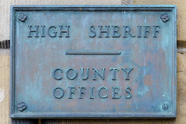 An antique weathered copper or brass sign with raised letters that say HIGH SHERIFF COUNTY OFFICES. The sign is on a building and tarnished with age..