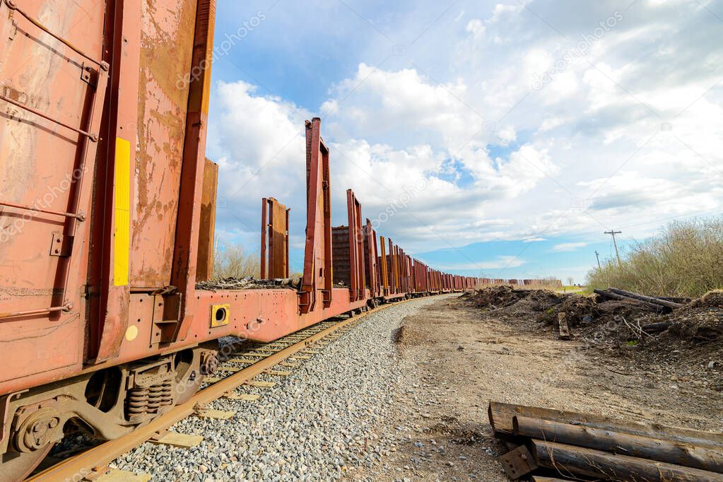 A long line of rail cars used to carry logs. Cars are empty. Cars curve right in the distance. Overcast sky but blue in the distance.