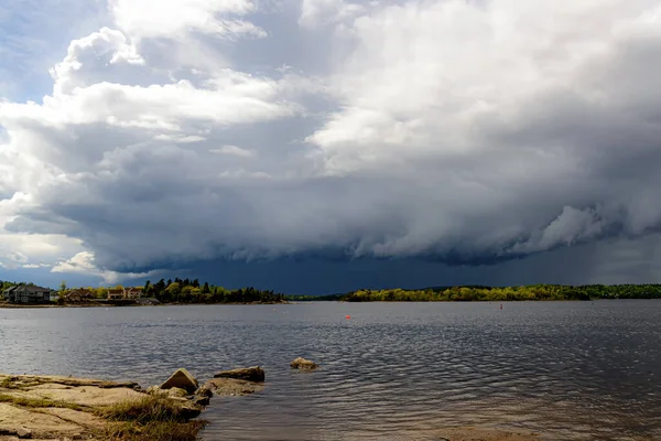 A large dark storm beneath dense clouds in the distance. It is dark blue gray under the clouds. Bright sky above.Water in the foreground.