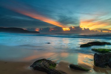 Ufuktaki bir bulut kümesinin üzerinde yüksek bulutlar sürükleniyor, gökyüzünde güneş ışınları yayılıyor, Orta Sahildeki Killcare Sahili 'nden güzel bir gündoğumu deniz manzarası yaratıyor, NSW, Avustralya.