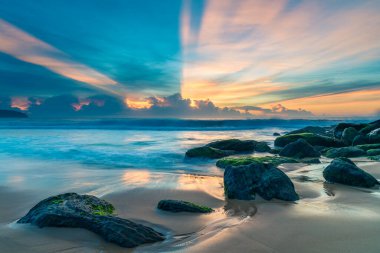 Ufuktaki bir bulut kümesinin üzerinde yüksek bulutlar sürükleniyor, gökyüzünde güneş ışınları yayılıyor, Orta Sahildeki Killcare Sahili 'nden güzel bir gündoğumu deniz manzarası yaratıyor, NSW, Avustralya.