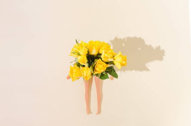 Women's legs emerging from a bouquet of bright yellow roses, on a beige background. Vibrant summer relaxation concept. Creative feminine floral design.