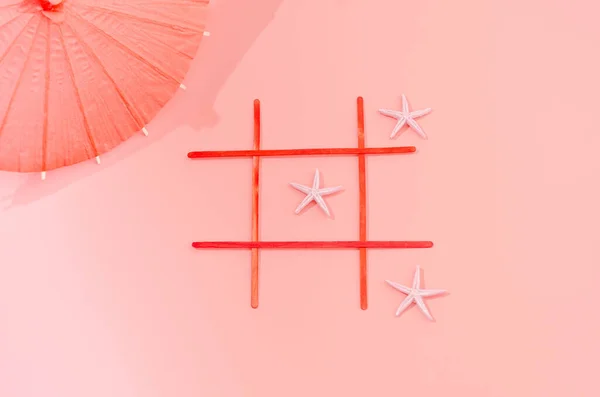A tic tac toe game with light pink stars and an orange umbrella on a salmon background. Minimal summer holidays design.