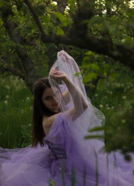 Ormanda bir kız ağaçların arasında çimenlerde oturur ve kumaşın içinden bakar.