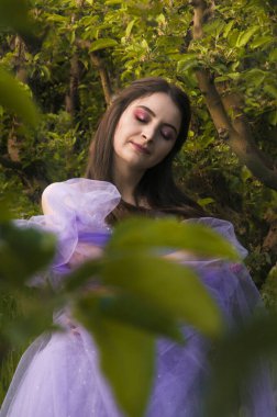 Ağaçların arasında, parlak makyajlı, gür elbiseli, gözleri kapalı bir kız.