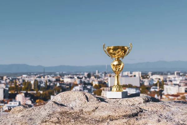 Gold trophy cup - aword for winner on high hill against background of city and blue sky. Achievement, success, victory, triumph concept. Copy space for text