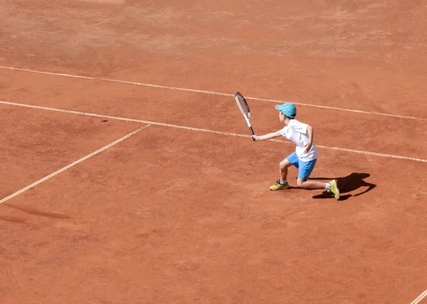Child tennis player on a clay court. A boy plays tennis on an outdoor tennis court. Kids tournament. The development of determination, strength and perseverance. Active sport. Copy space.
