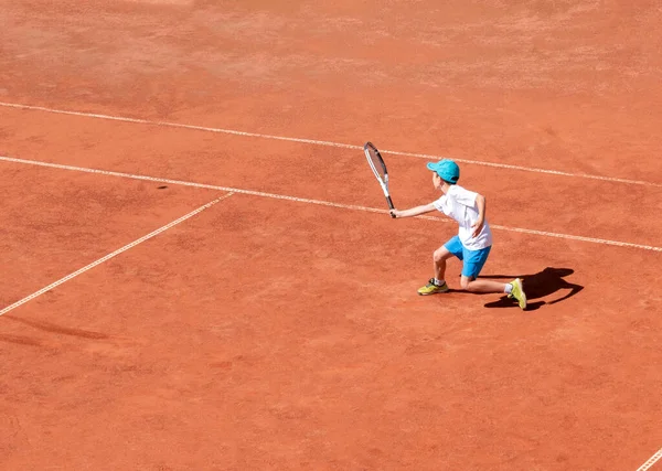 Child tennis player on a clay court. A boy plays tennis on an outdoor tennis court. Kids tournament. The development of determination, strength and perseverance. Active sport. Copy space.