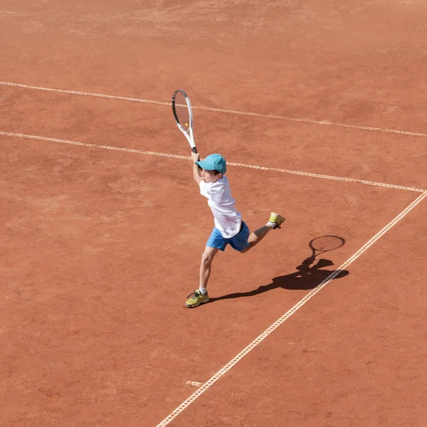 A boy plays tennis on a clay court. A little tennis player focused on the game and shot in flight after hitting the ball. Tennis kid. Sports action frame. Active games. Square size.