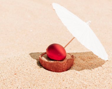 Kırmızı Paskalya yumurtası hindistan cevizi kabuğunda güneş şemsiyesi altında kumlu kumsalda. Seyahat, tatil, tropik ülkelerin sahillerindeki sahil beldesinde Paskalya tatili. Paskalya arkaplanı, Tebrik kartı kopyalama alanı