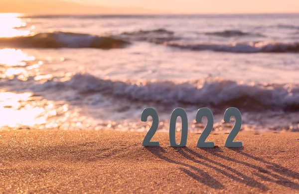 Mutlu yıllar 2022. Günbatımında deniz tropikal kumsalının altın kumu üzerinde 2022 numaranın sembolü. Bulanık arka planda dalgalar hareket ediyor. Yeni yıl kartını selamlıyorum. Seçici odaklanma. Boşluğu kopyala