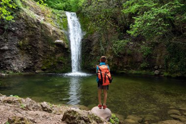 Çocuk çocuk, küçük bir yürüyüşçü sırt çantasıyla düşen şelale suyuna hayran. Doğa terapisi, yürüyüş, konaklama, yerel seyahat, aile gezisi konsepti. Rodop Dağları, Bulgaristan