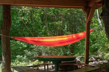 Orange hammock is stretched in green forest near picnic site with wooden table and benches. Summer vacation, hiking, local tourism, eco travel, resting and relaxation among nature outdoors concept