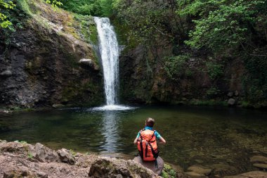 Child boy schoolboy with hiking backpack sitting alone in forest near lake and watching waterfall. Children's exploration and curiosity of nature, adventure learning and outdoor education