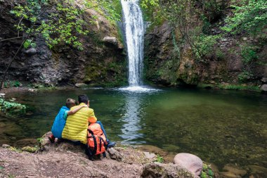 Son hugging dad, sitting near lake looking falling stream of water of waterfall, happy together among nature. Family vacancy, hiking, child and father love. Back view. Father's day concept