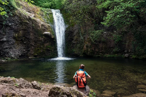 Child boy schoolboy with hiking backpack sitting alone in forest near lake and watching waterfall. Children's exploration and curiosity of nature, adventure learning and outdoor education
