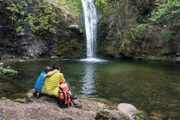 Son hugging dad, sitting near lake looking falling stream of water of waterfall, happy together among nature. Family vacancy, hiking, child and father love. Back view. Father's day concept