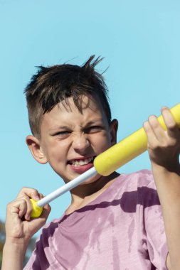 Close-up portrait child boy playing with water gun on hot summer day, shoots with toy pistol against background of blue sky. Summer children's games and fun, water war