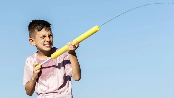 Kids summer game, water war. Close-up portrait child boy playing with water gun on hot summer day, shoots with toy pistol against background of blue sky. Baner, copy space