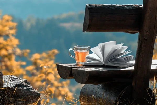 Sonbahar açık hava eğlence sahnesi. Açık kitap ve cam şeffaf fincanda bitki çayı, sonbahar ormanlarının arka planı ve sonbahar renklerinde dağlar. Okuma, rahatlama, yalnızlık, doğada eğlence.