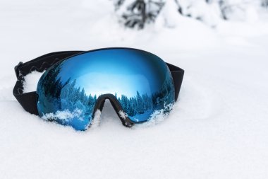 Taze kar üzerinde duran mavi aynalı mercekli kayak gözlükleri, kış ormanı ve dağları yansıtıyor, kış sporlarını ve macerayı sembolize ediyor, Noel tatilleri ve kayak tatili kavramını.