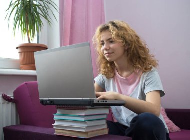 Güzel kız - 17 yaşında bir genç evinde sınavlara hazırlanıyor, bir kanepede dizüstü bilgisayarla oturuyor. Evde eğitim, sosyal ağlar, uzaktan öğrenme. Bilgi, kitaplar, modern dijital teknolojiler