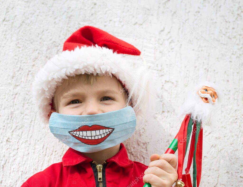 kid in Santa hat . close up portrait.  On his face there is a blue medical mask with painted funny smile. Time for gifts. Stay home. Wear masks. Positive thinking. Christmas Eve