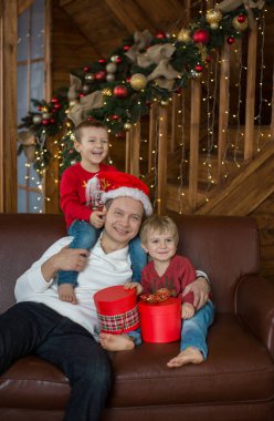 Noel Baba şapkalı bir baba ve çıplak ayaklı oğulları 4 yıl boyunca kırmızı kazaklar içinde güzel dekore edilmiş yeni yıl evinde hediye kutuları ile. Herkes neşeyle gülümsüyor, sevinç ve kutlama duyguları. Noel Arifesi