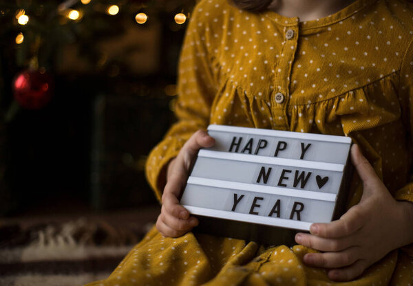 Крупным планом девушки в горчичном платье с табличкой "С Новым годом". Боке на темном фоне, место для текста, копировальное пространство