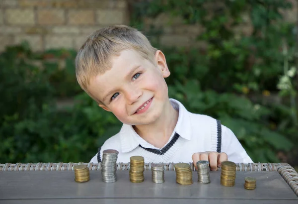 Portrait of a little cute boy in front of whom on the table are lined with columns of coins. focus on the face. Little financier, investment in the future. Child collects money for a dream