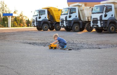 cute toddler boy interestedly plays with toy dump truck on control panel sitting on asphalt. behind him are 3 large real dump trucks. sunny warm day. Favorite games for boys, happy childhood