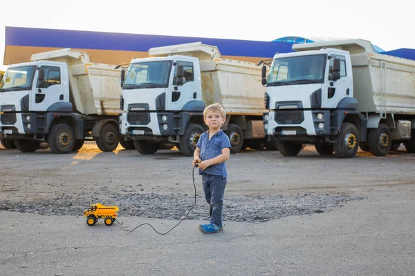 cute boy plays with a toy dump truck on control panel on asphalt. behind him are large real dump trucks. sunny warm day. Favorite games for boys